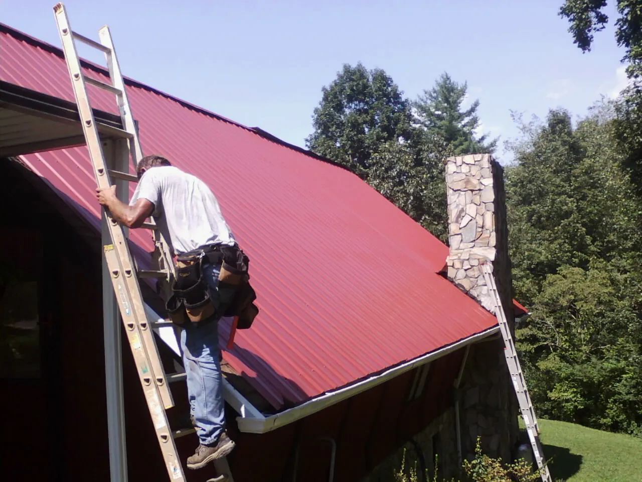 Roofer on a ladder beside a red metal roof and stone chimney — Soffit & Fascia Repair by Roofing Brothers in South Dartmouth