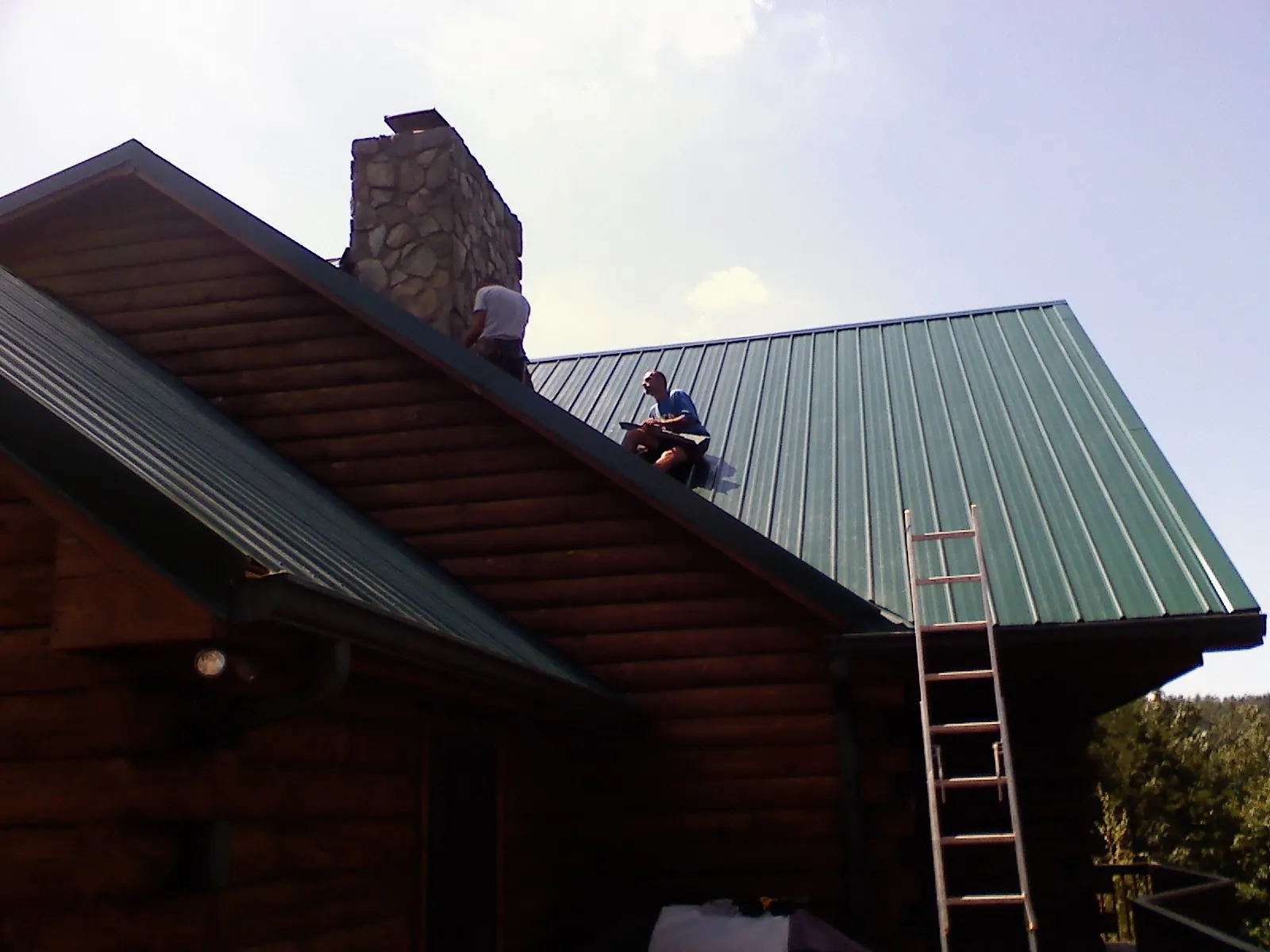 Roofers installing green metal panels beside a stone chimney on a log home in South Dartmouth